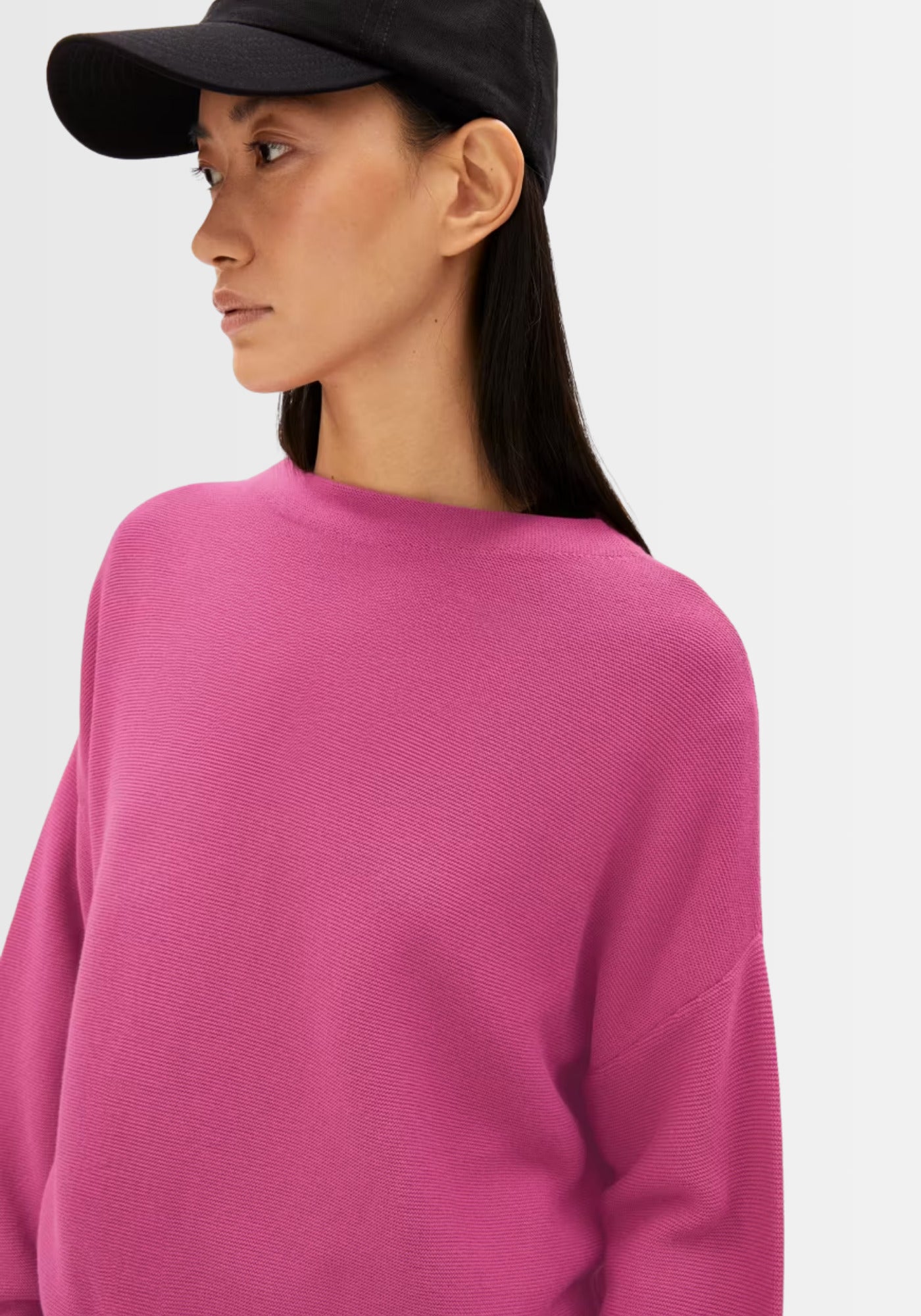 Model trägt den lockeren Merinaa Links Links Feinstrickpullover in kräftigem Pink, kombiniert mit schwarzer Cap und Jeans, ideal für einen sportlich-modernen Stil.