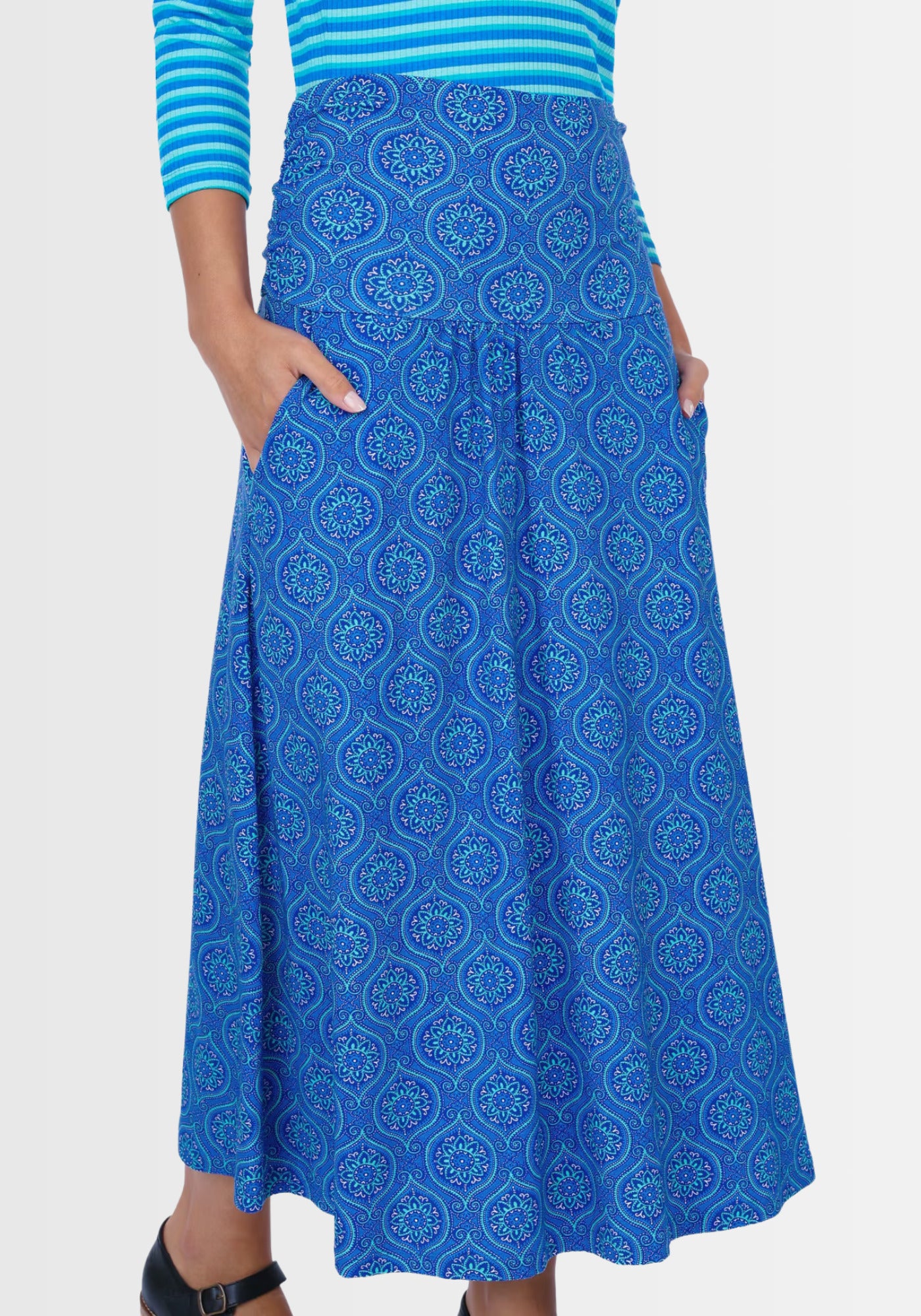 Ein Model kombiniert den Rock „Kalla“ in Lapiston mit einem schlichten Shirt und Sandalen, um einen boho-inspirierten Sommerlook zu kreieren. Die lebhaften Ornamente, die praktischen Taschen und das elastische Bündchen verleihen Style und Funktionalität.